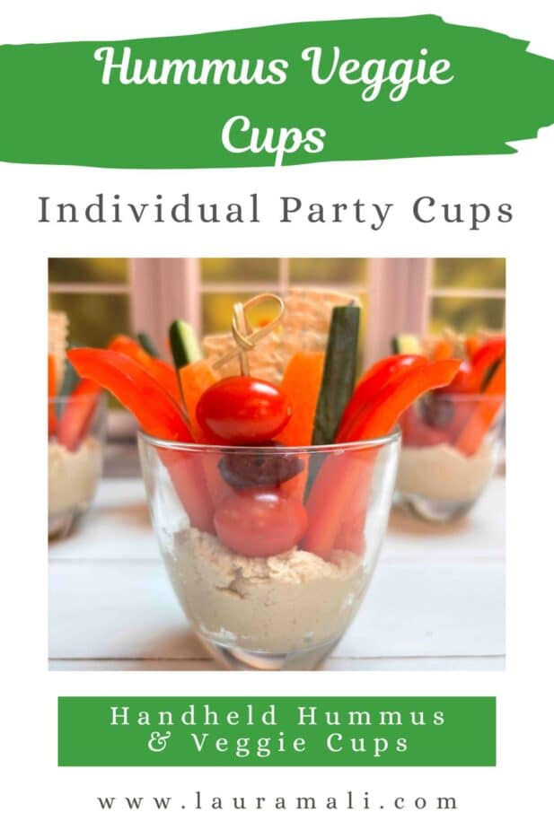 A glass cup with the bottom filled with hummus and a variety of veggie sticks and crackers dipped in the hummus and poking out the top of the glass. Additional cups are in the background. 