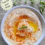 A white bowl filled with creamy hummus on a wood table. The hummus is garnished with a sprinkle of smoked paprika and a sprig of fresh oregano.