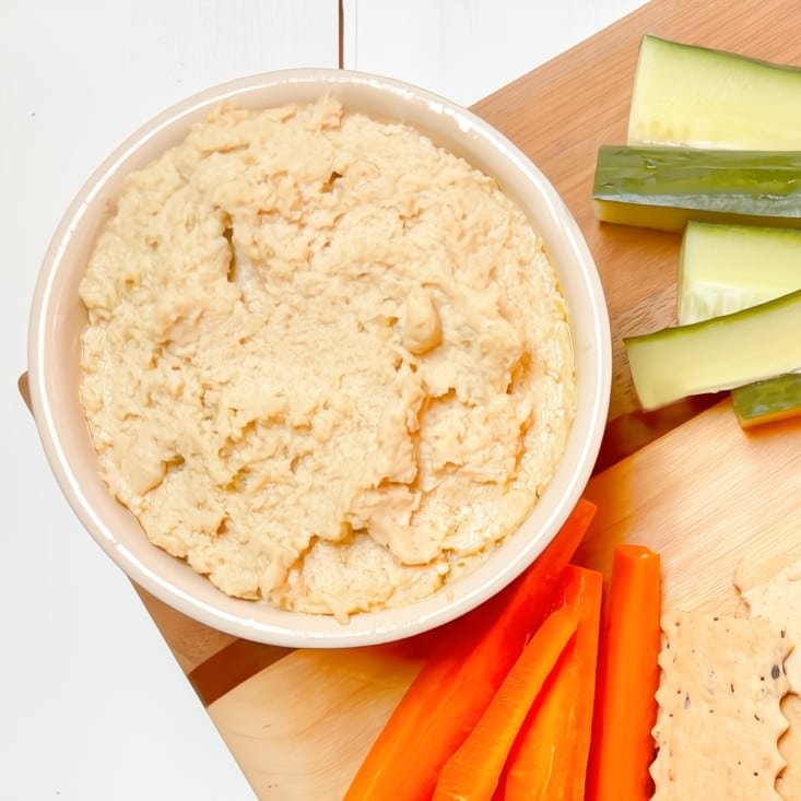 A bowl of hummus with cut up veggies and crackers on a wooden board