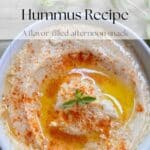 A white bowl filled with creamy hummus on a wood table. The hummus is garnished with a sprinkle of smoked paprika and a sprig of fresh oregano.