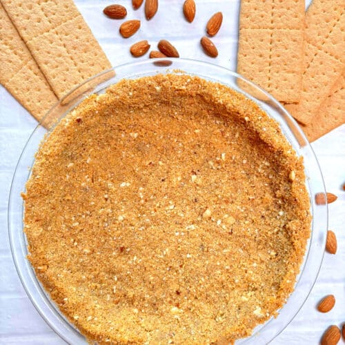 A graham cracker pie crust in a glass pie pan surrounded by graham crackers and almonds.