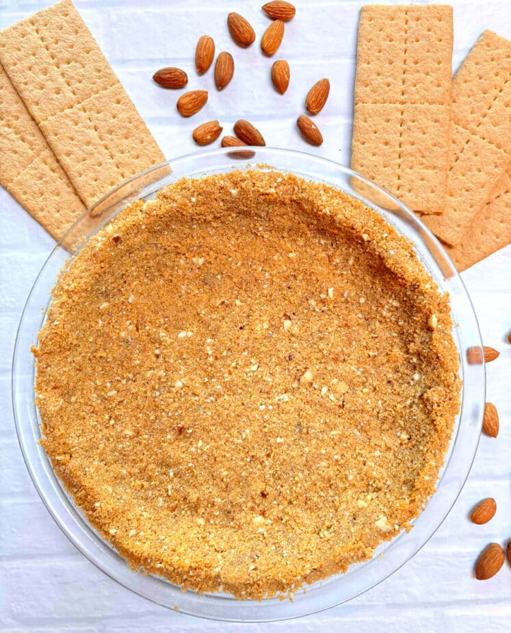 A graham cracker pie crust in a glass pie pan surrounded by graham crackers and almonds.