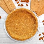 A baked graham cracker pie crust on a white counter with graham crackers and almonds surrounding it.