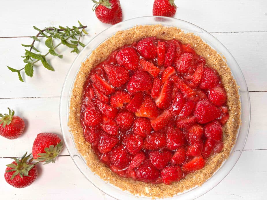 Strawberry Pie in a Graham Cracker Crust on a white picnic table with strawberries and mint surrounding it. 