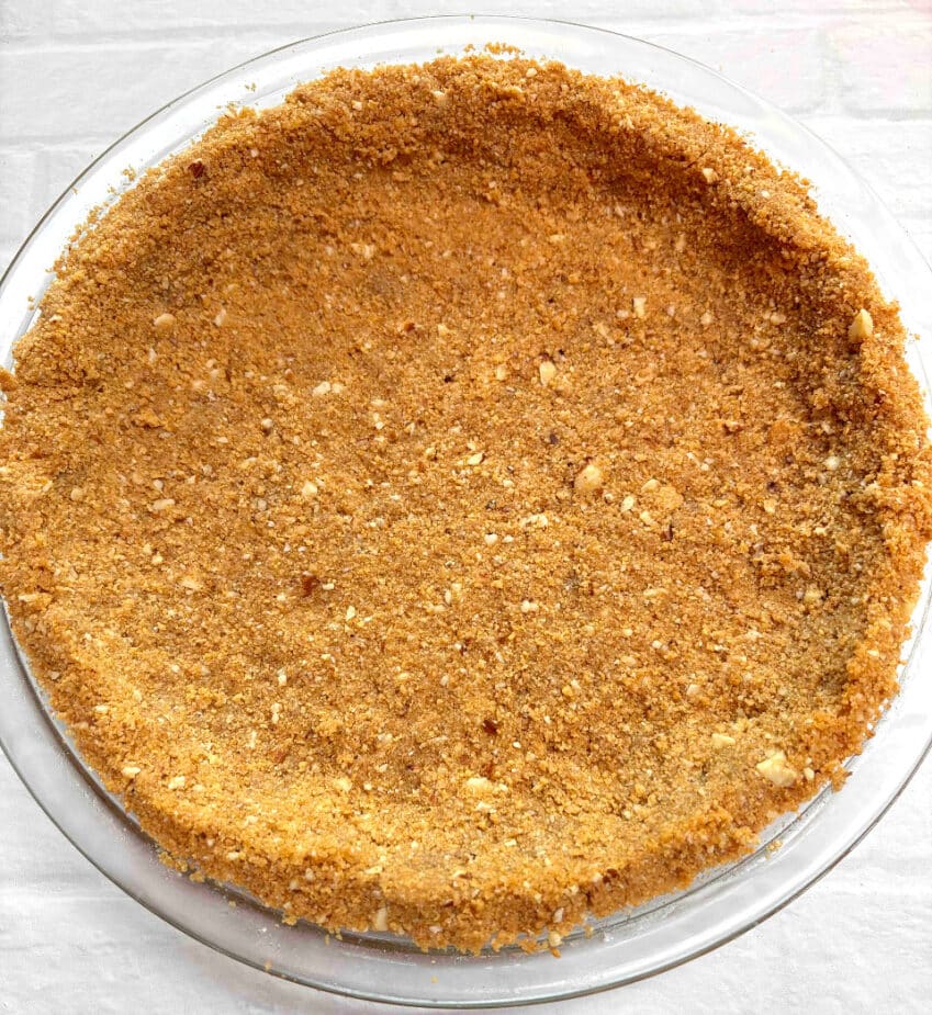 A baked graham cracker pie crust in a glass pie dish on a white brick counter. 
