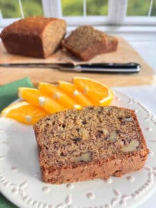 A slice of banana bread on a white plate with orange slices. A cut loaf of bread is in the background