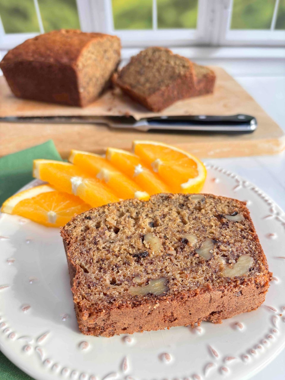 A slice of banana bread on a white plate with orange slices. A cut loaf of bread is in the background