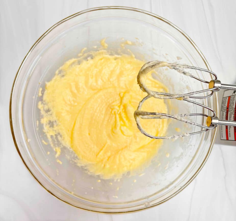 Butter, sugar and eggs beaten together in a glass bowl. The mixer is on the right side of the bowl with the beaters over top. 