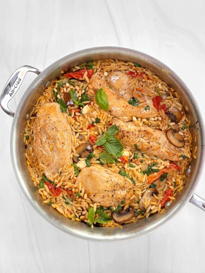 A saute pan filled with cooked orzo, chicken breasts, sundried tomatoes, mushrooms and spinach garnished with fresh basil and pine nuts. It is sitting on a marble counter.