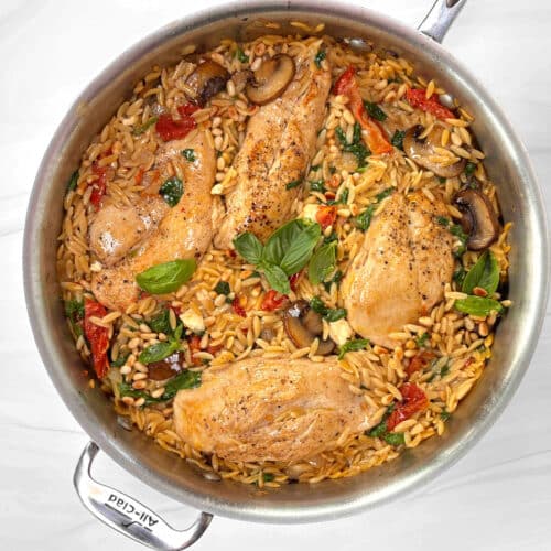 A sautepan filled with orzo, sundried tomatoes, mushrooms and chicken breasts on a marble counter