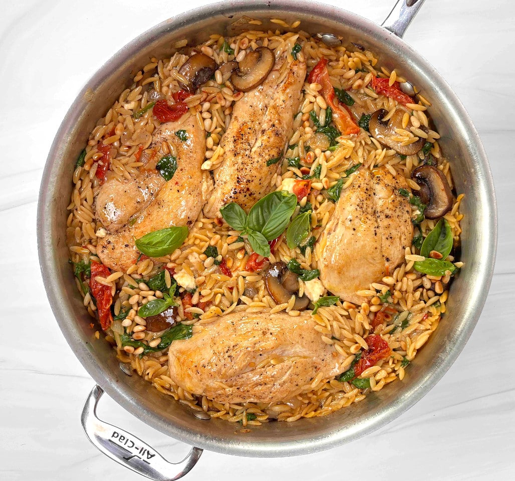 A sautepan filled with orzo, sundried tomatoes, mushrooms and chicken breasts on a marble counter