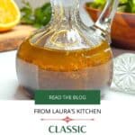 A small glass carafe filled with orange balsamic vinaigrette salad dressing on a white counter. A salad and cut orange are in the background.