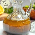 A small glass carafe filled with orange balsamic vinaigrette salad dressing on a white counter. A salad and cut orange are in the background.