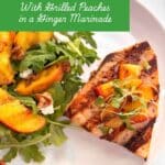 A swordfish steak on a white plate with grill marks. It is topped with grilled peaches. An argula salad with grilled peaches is on the side. The title "Grilled Swordfish with grelled peaches in a ginger marinade is in a green box on the top.