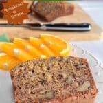 A slice of banana bread on a white plate with orange slices. A loaf of cut bread on a wood cutting board is in the background with a knife. "Easy Spiced Banana Bread" is in a green banner at the top