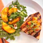 A white plate with a grilled swordfish steak topped with grilled peaches. On the side is a salad made of arugula and goat cheese topped with extra grilled peaches. A peach colored napkin and fork are on the side.