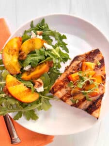 A grilled swordfish steak topped with grilled peaches on a white plate with a salad made with arugula and topped with extra grilled peaches on the side.