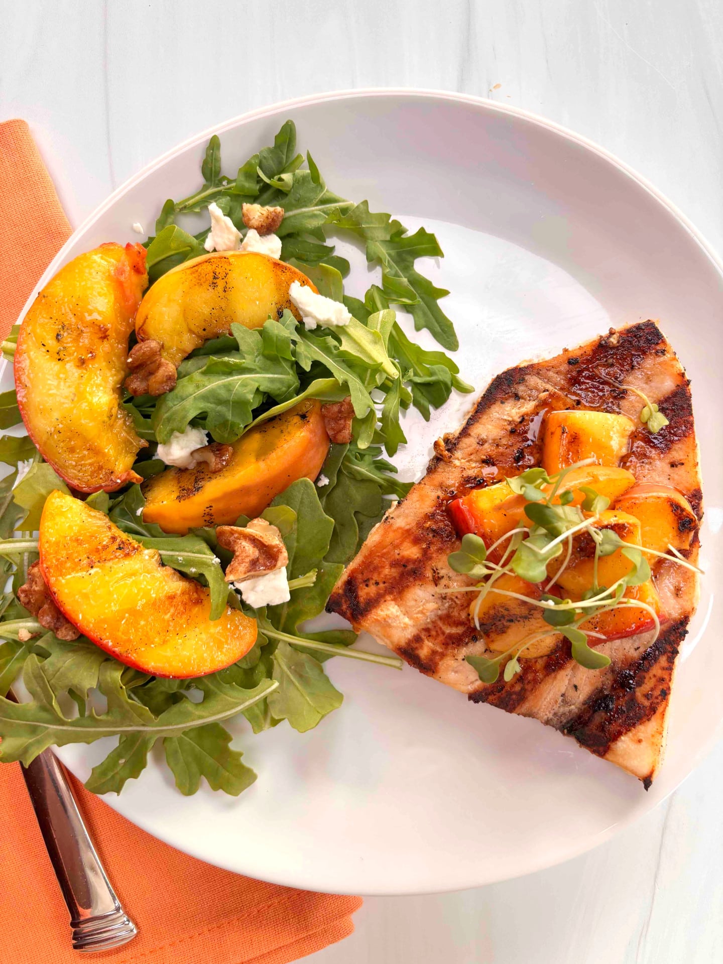 A grilled swordfish steak topped with grilled peaches on a white plate with a salad made with arugula and topped with extra grilled peaches on the side.