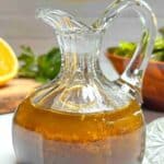A glass carafe filled with the orange balsamic vinaigrette dressing on a white counter. A green salad and half orange are in the background.
