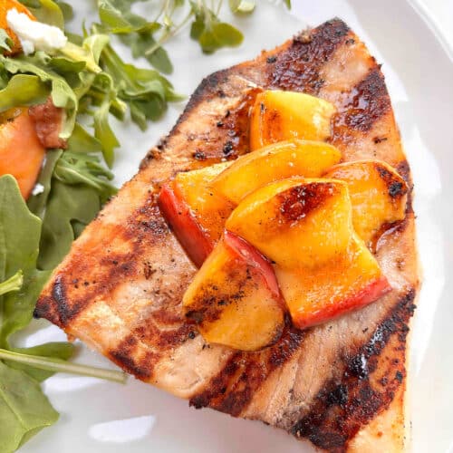 A grilled swordfish steak on a white plate, topped with grilled peaches and an arugula salad is next to it on the white plate.