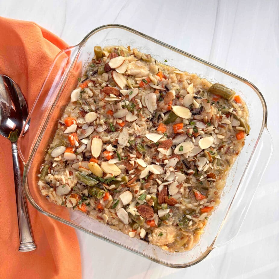 A square glass casserole dish filled with wild rice, salmon and vegetables that is topped with almonds. An orange linen and spoon are on the side.
