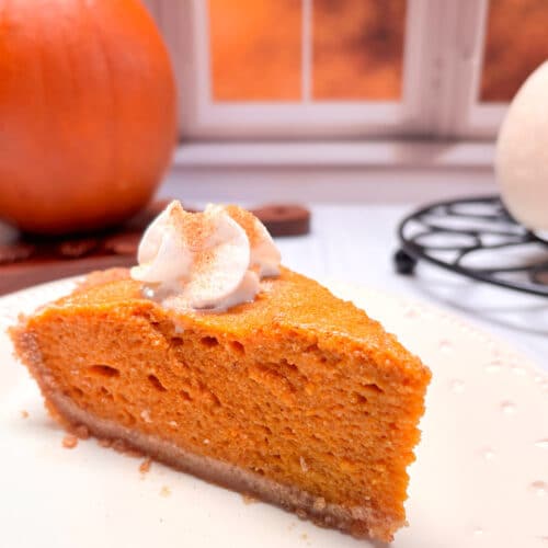 A piece of pumpkin chiffon pie on a white plate. A pumpkin is in the background next to a window with a fall scene.
