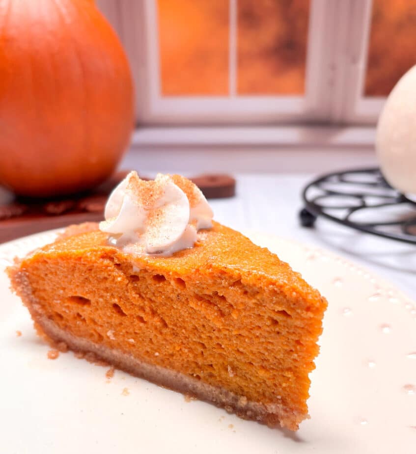 A piece of pumpkin chiffon pie on a white plate. A pumpkin is in the background next to a window with a fall scene.