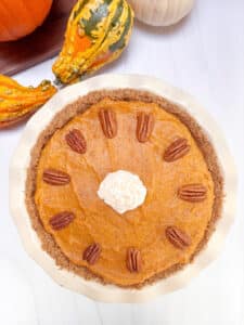A pumpkin chiffon pie in a pie dish on a white marble counter. The pie is decorated with pecans and whipped cream. 2 fall gourds are in the background.