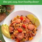 A white bowl filled with rice, vegetables and slices of turkey kielbasa with sliced avocado and limes on the side.