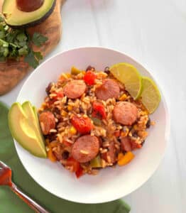 A white bowl filled with Southwestern Turkey Sausage, Rice, and vegetables. Slices of avocado and limes are on the side.