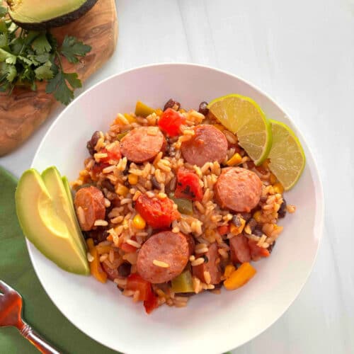 A white bowl filled with Southwestern Turkey Sausage, Rice, and vegetables. Slices of avocado and limes are on the side.