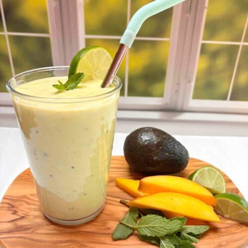 A mango smoothie on a wood cutting board with an avocado, mango slices, mint and lime next to it.