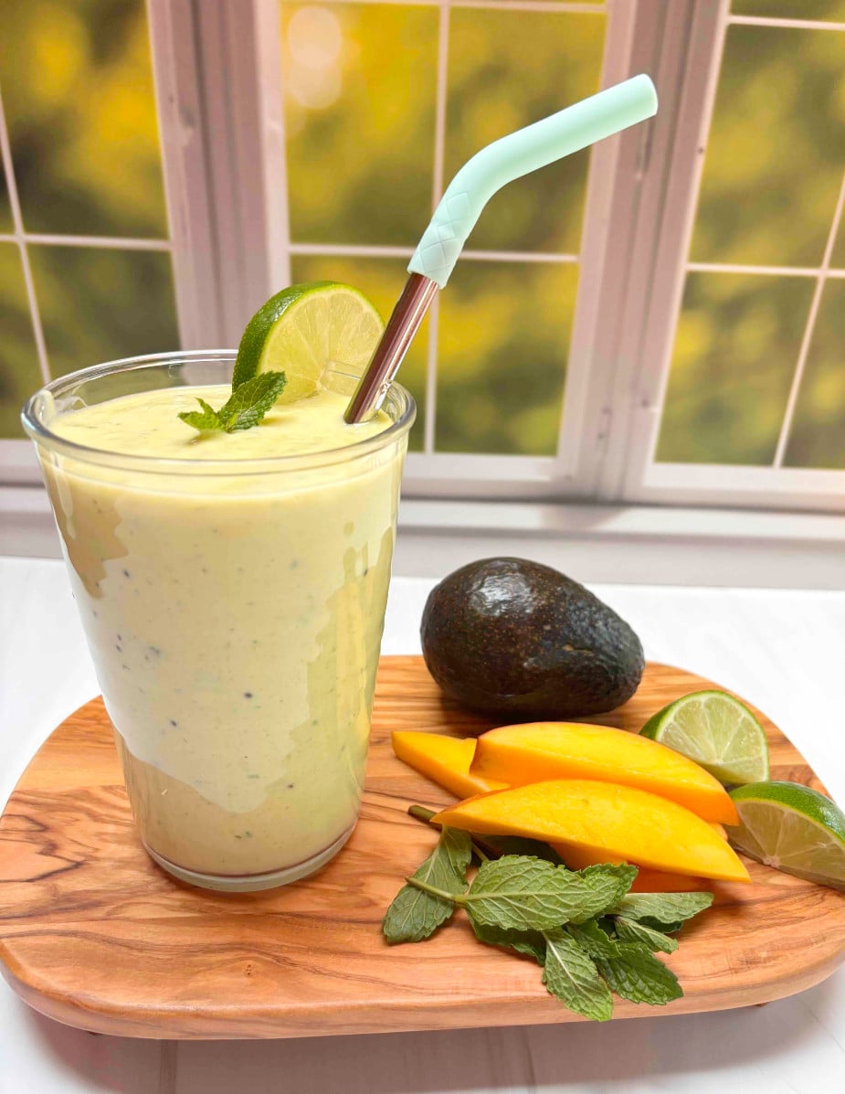 A mango smoothie on a wood cutting board with an avocado, mango slices, mint and lime next to it.