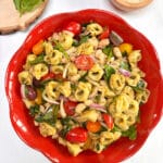 A round red bowl filled with tortellini pasta salad with fresh basil and parmesan cheese in the background