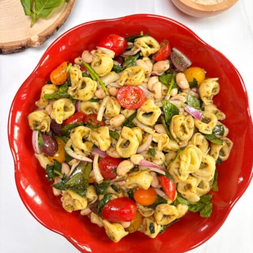 A round red bowl filled with tortellini pasta salad with fresh basil and parmesan cheese in the background