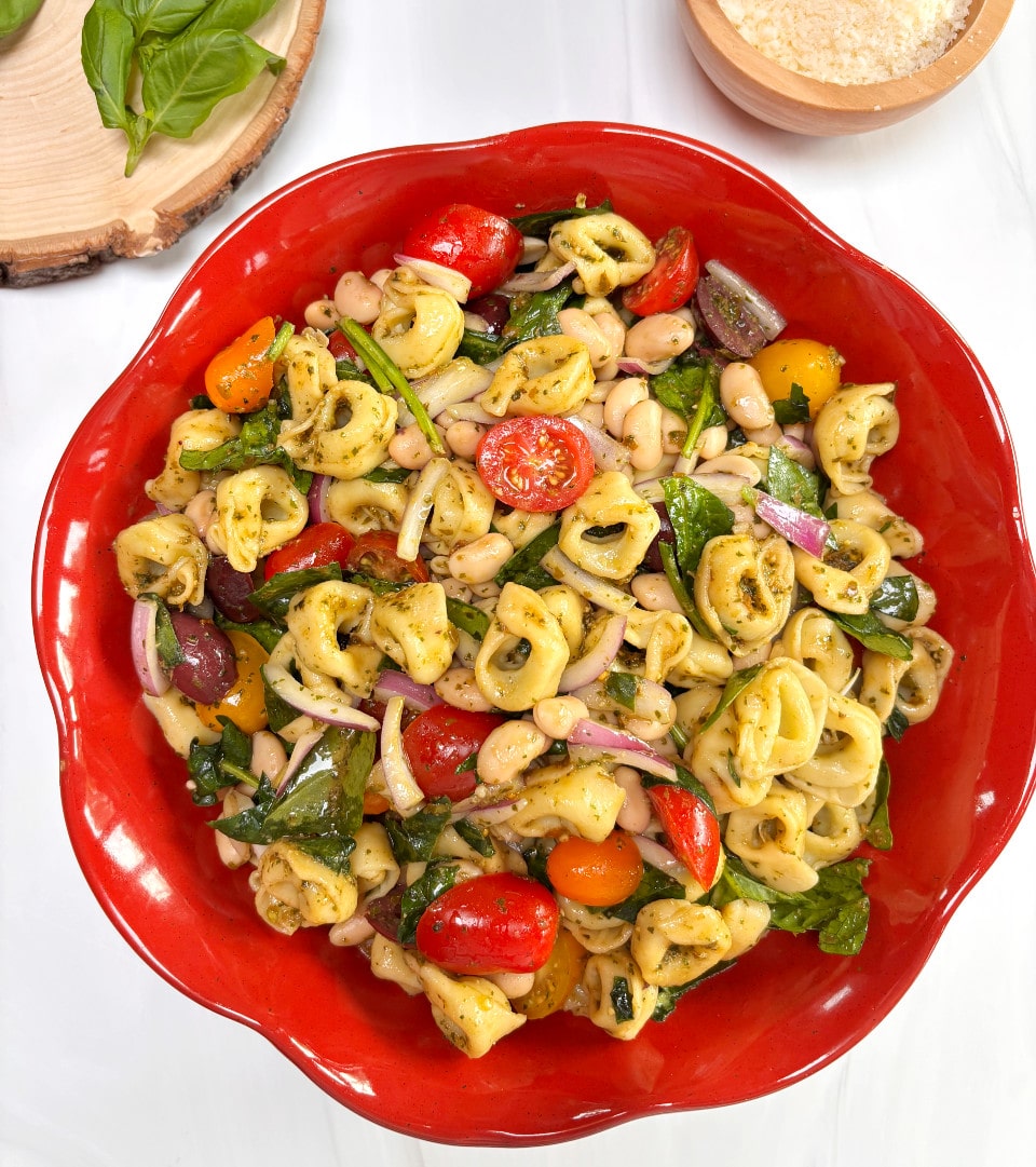 A round red bowl filled with tortellini pasta salad with fresh basil and parmesan cheese in the background