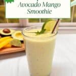A mango smoothie on a white table overlooking a bright green yard. An avocado, lime and mango slices are in the background on a cutting board.