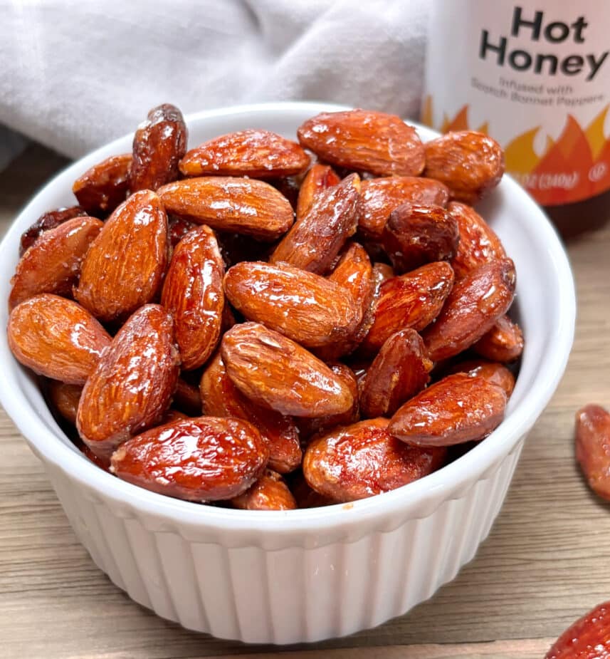 Hot Honey Almonds A small white bowl filled with hot honey roasted almonds with a container of "hot honey" in the background