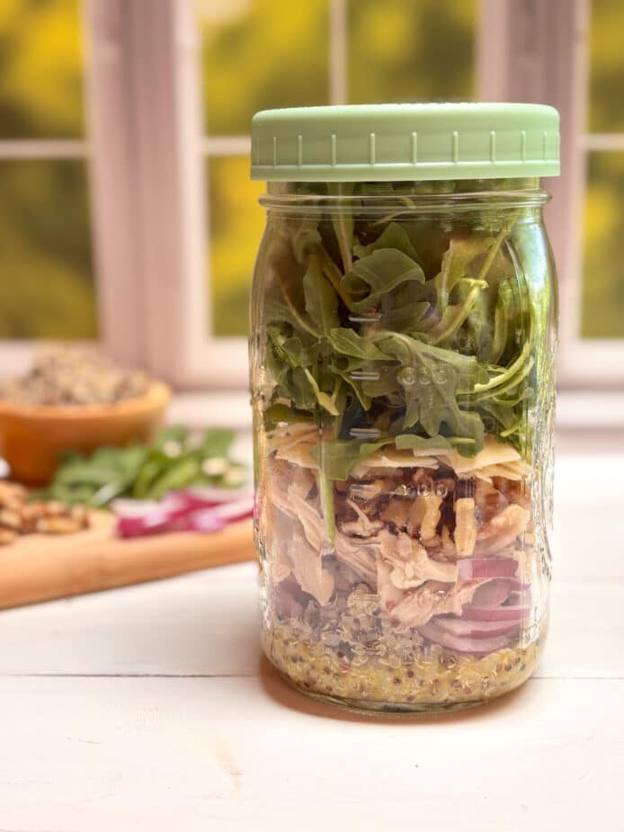 Jar Lemon Arugula Salad A mason jar with layered ingredients for the Lemon Arugula Salad. A cutting board with the ingredients is in the background.
