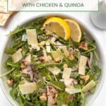 A white bowl filled with Arugula, chicken, quinoa, onions, walnuts, and shaved parmesan cheese with a cruet of dressing in the the background.