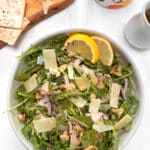 A bowl of arugula with chicken, shredded parmesan cheese, walnuts, quinoa and onions. A cruet of dressing with crackers and walnuts in the background.