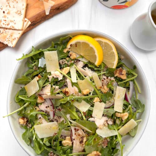 A bowl of arugula with chicken, shredded parmesan cheese, walnuts, quinoa and onions. A cruet of dressing with crackers and walnuts in the background.