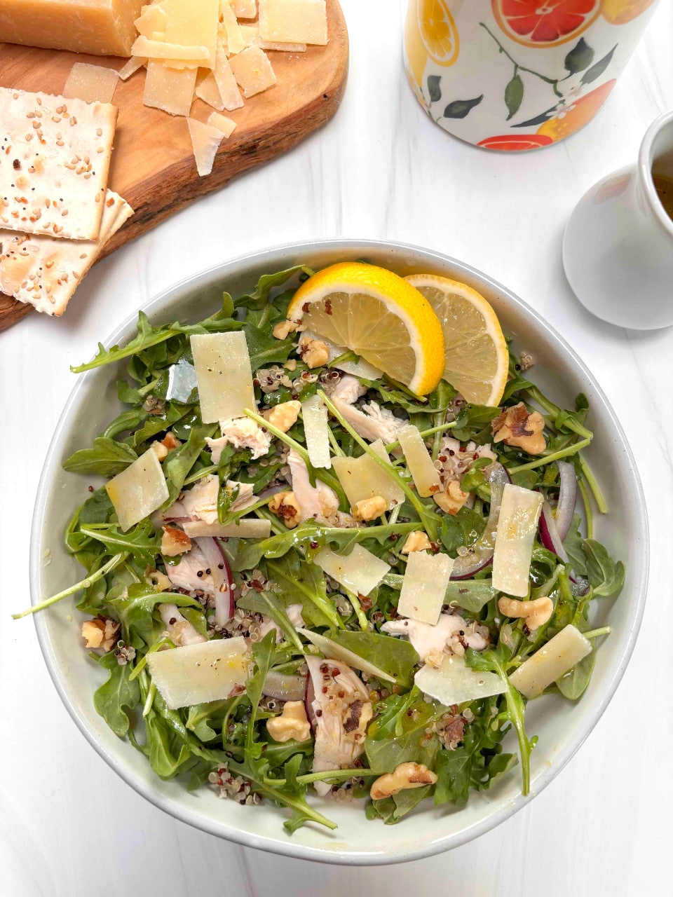 A bowl of arugula with chicken, shredded parmesan cheese, walnuts, quinoa and onions. A cruet of dressing with crackers and walnuts in the background.