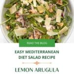 A close up of a bowl of arugula toped with chicken, shaved Parmesan, walntus and quinoa and lemon slices.