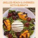 A green salad in a white bowl topped with grilled peaches and cherries and a ball of burrata cheese in the center.