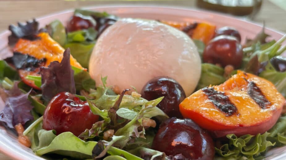 Close up Peach and Burrata A ball of burrata cheese sits on top of leafy greens surrounded by grilled peaches and cherries.
