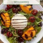 A white bowl filled with mixed greens, grilled peaches and cherries and burrata cheese drizzled with balsamic vinegar.