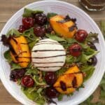 A bowl filled with salad greens topped with grilled peaches and cherries and a round burrata cheese ball in the center. A drizzle of balsamic is over top.