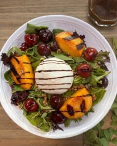 A bowl filled with salad greens topped with grilled peaches and cherries and a round burrata cheese ball in the center. A drizzle of balsamic is over top.