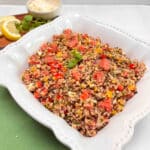 A white bowl filled with quinoa watermelon salad. A dish of feta cheese, mint and lemon slices are in the background cheese